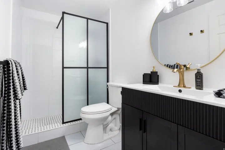 Modern bathroom featuring a black vanity with textured paneling, a gold-framed circular mirror, a gold-finish faucet, a white integrated sink, and a shower with a black-framed frosted glass door