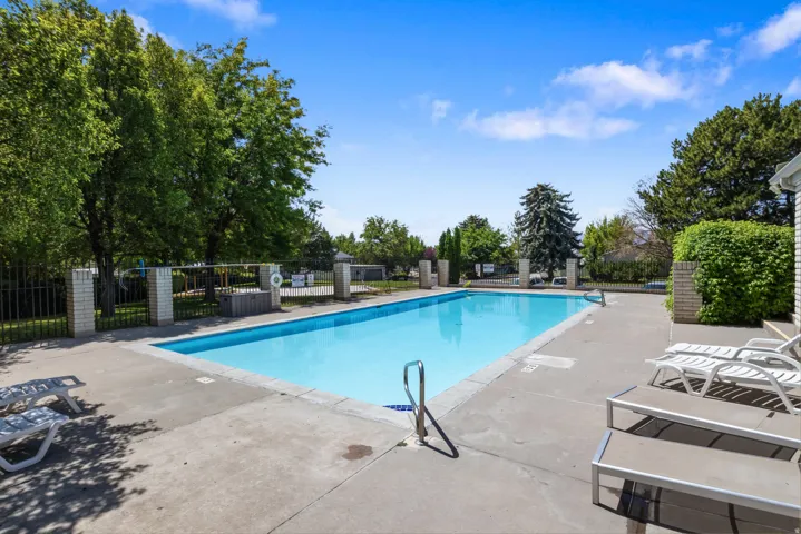 Outdoor swimming pool with concrete decking, surrounded by mature trees and a black metal fence with brick pillars