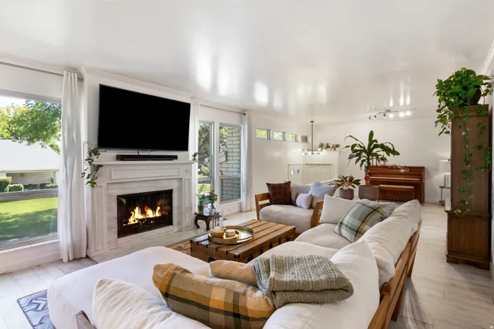 Living area with a fireplace featuring a white mantel, large windows, wood-finish flooring, and crown molding