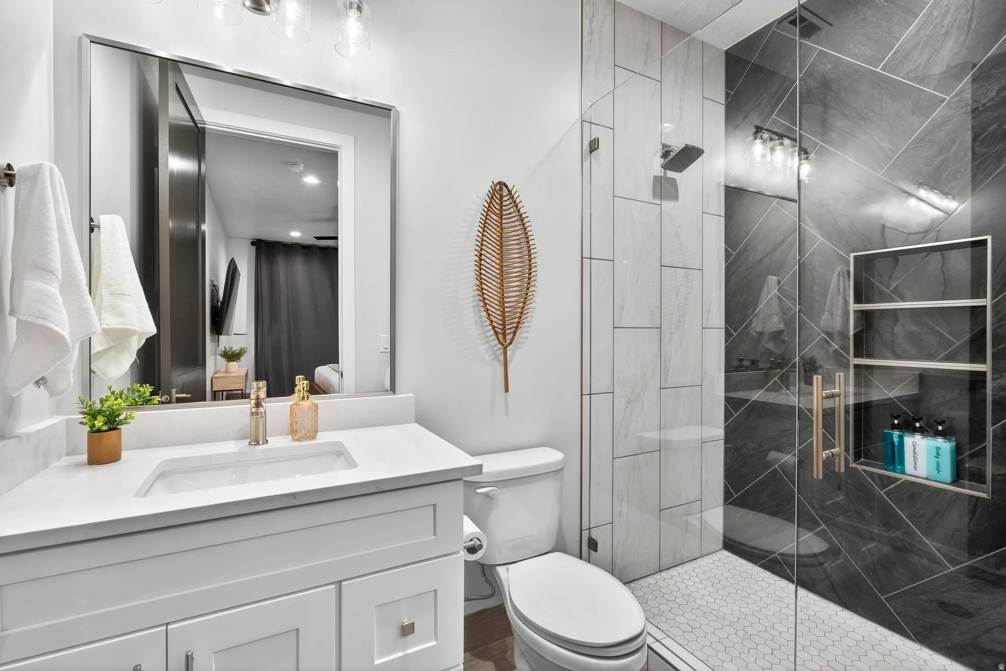 Modern bathroom featuring a white vanity with rectangular undermount sink and quartz countertop, large framed mirror with integrated lighting, walk-in shower with black tile surround and frameless glass enclosure, and woven pendant accent