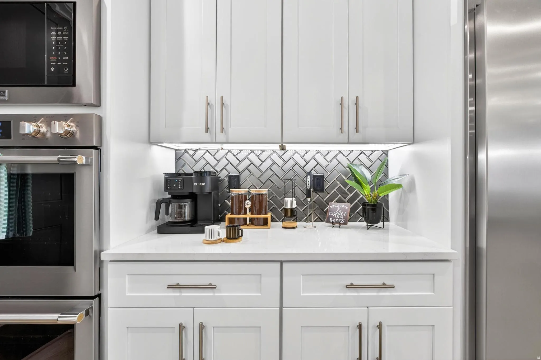 Integrated stainless steel microwave and oven, white shaker-style cabinetry, light-toned countertop, and a herringbone pattern tile backsplash