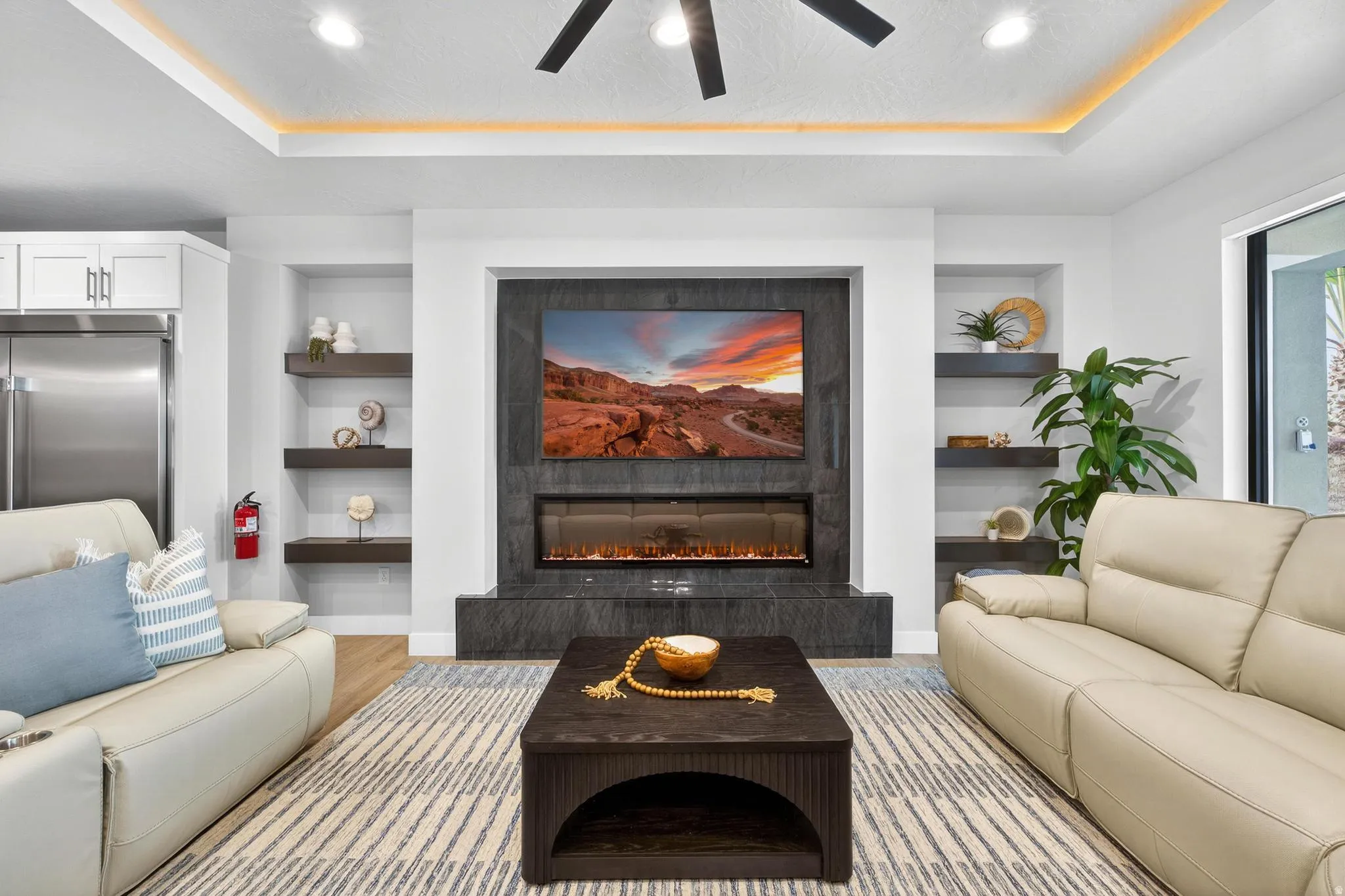 Recessed ceiling with perimeter lighting, ceiling fan, built-in shelving, stone-clad fireplace, and wood-finish flooring