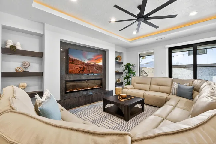 Recessed ceiling with accent lighting, large black ceiling fan, built-in shelving, and a wall-mounted linear fireplace