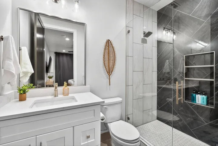 Modern bathroom featuring a white vanity with rectangular undermount sink and quartz countertop, large framed mirror with integrated lighting, walk-in shower with black tile surround and frameless glass enclosure, and woven pendant accent
