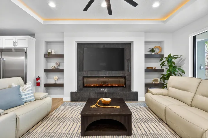 Coffered ceiling with accent lighting, ceiling fan, built-in shelving, and a wall-mounted electric fireplace with stone-finish surround