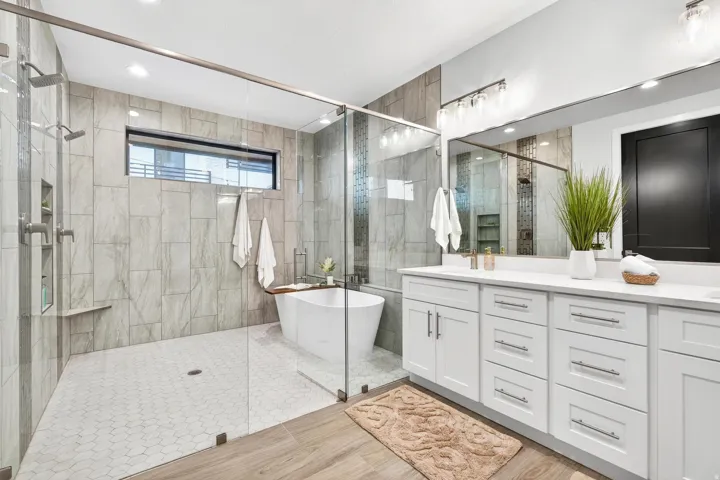 Spacious bathroom featuring a frameless glass shower enclosure, freestanding soaking tub, dual vanity with white cabinetry, and large format wall tiling
