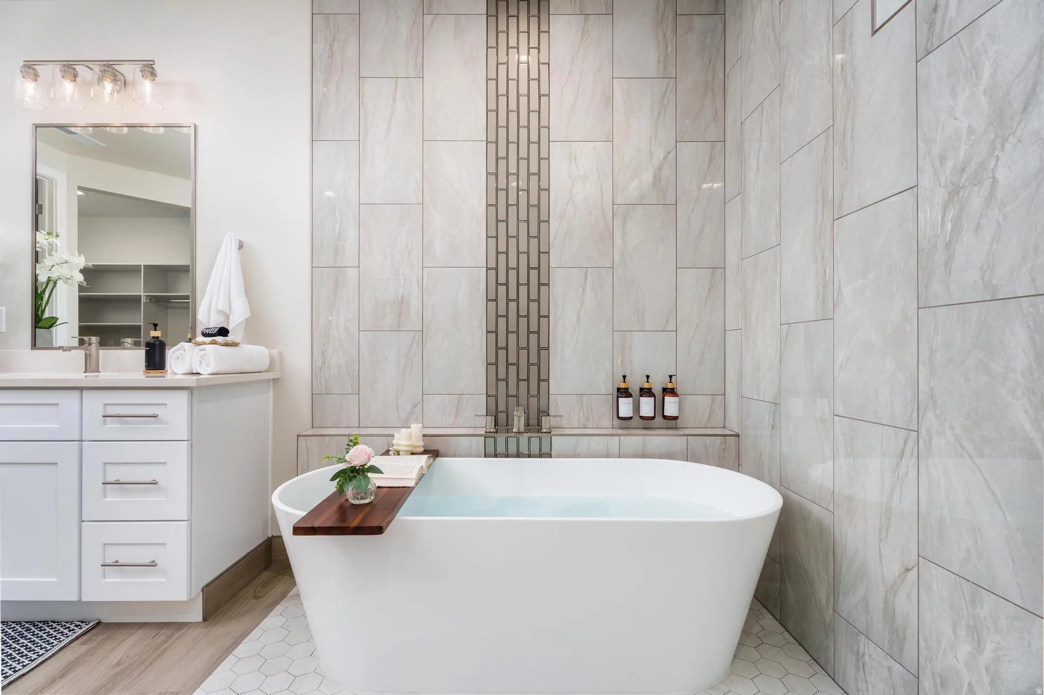 Freestanding soaking tub, white shaker vanity with drawer storage, polished chrome hardware, quartz countertop, and large format wall tiling