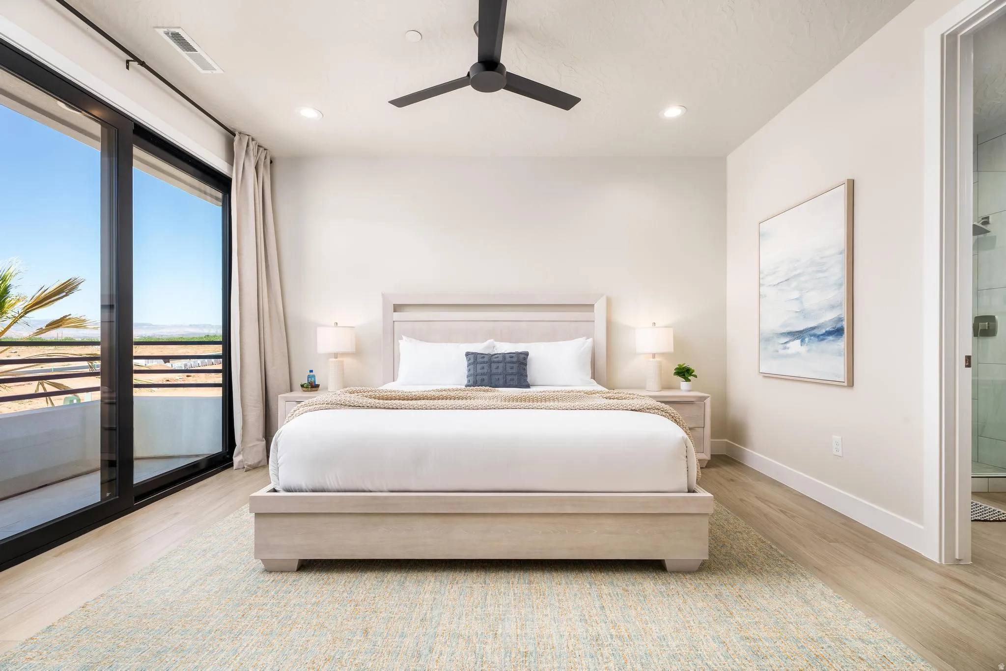 Full-height sliding glass doors, light wood-finish flooring, a contemporary ceiling fan, recessed lighting, and an adjacent full bathroom featuring a glass-enclosed shower