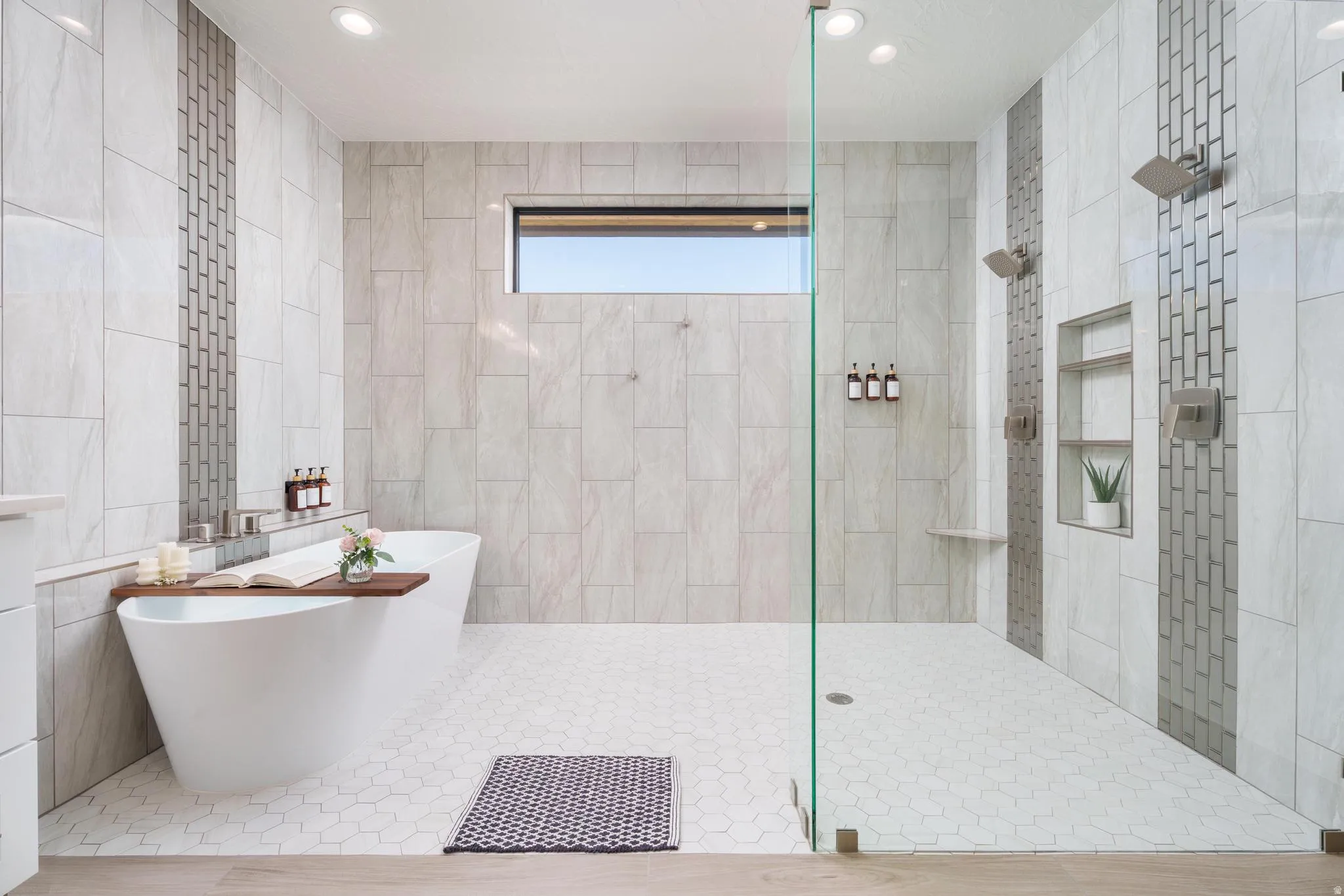 Freestanding soaking tub, walk-in shower with glass enclosure, dual shower heads, recessed shelving, and a horizontal window