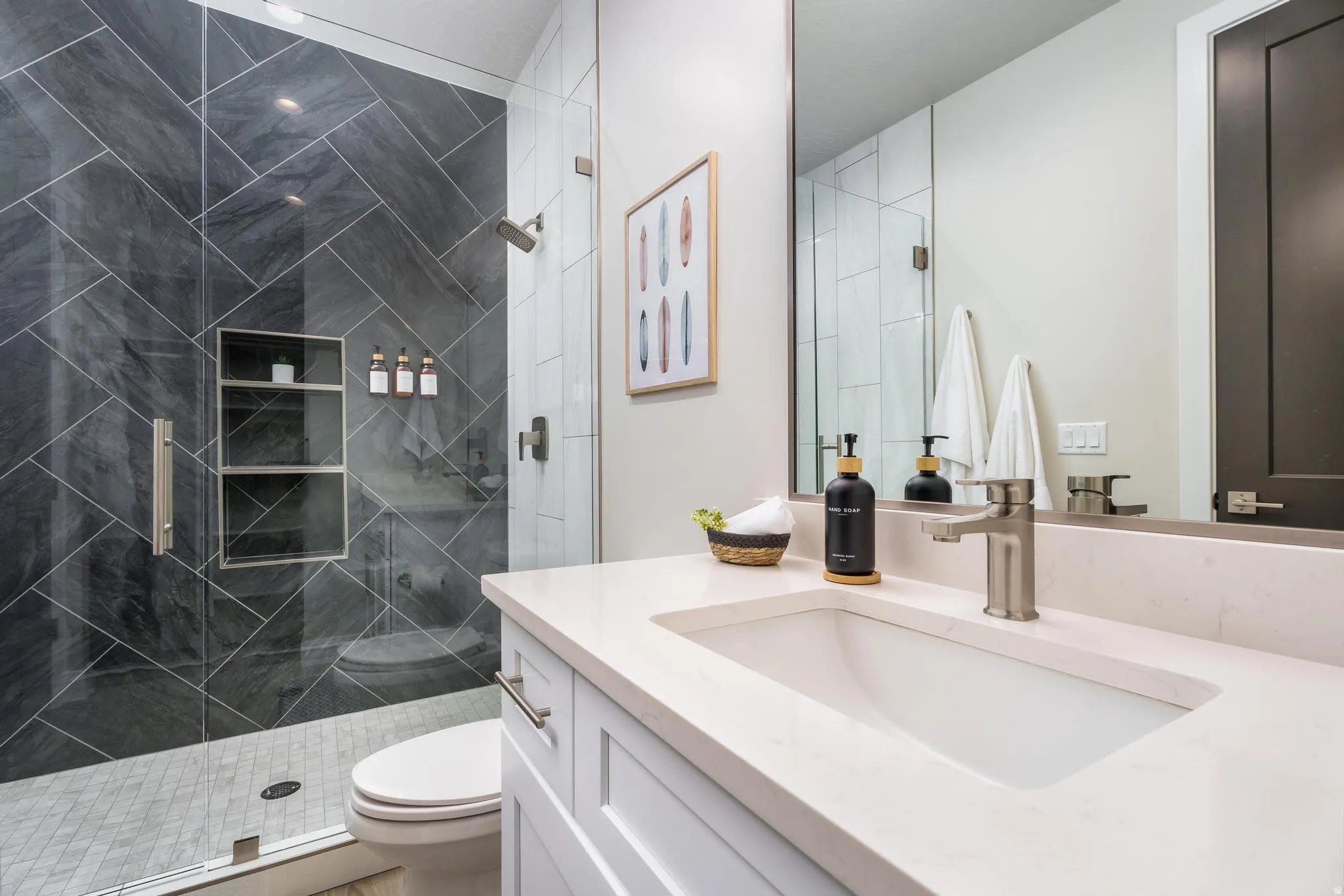 Glass-enclosed shower with dark gray diagonal tile and built-in shelving, white integrated sink, light-colored countertop, and brushed nickel fixtures