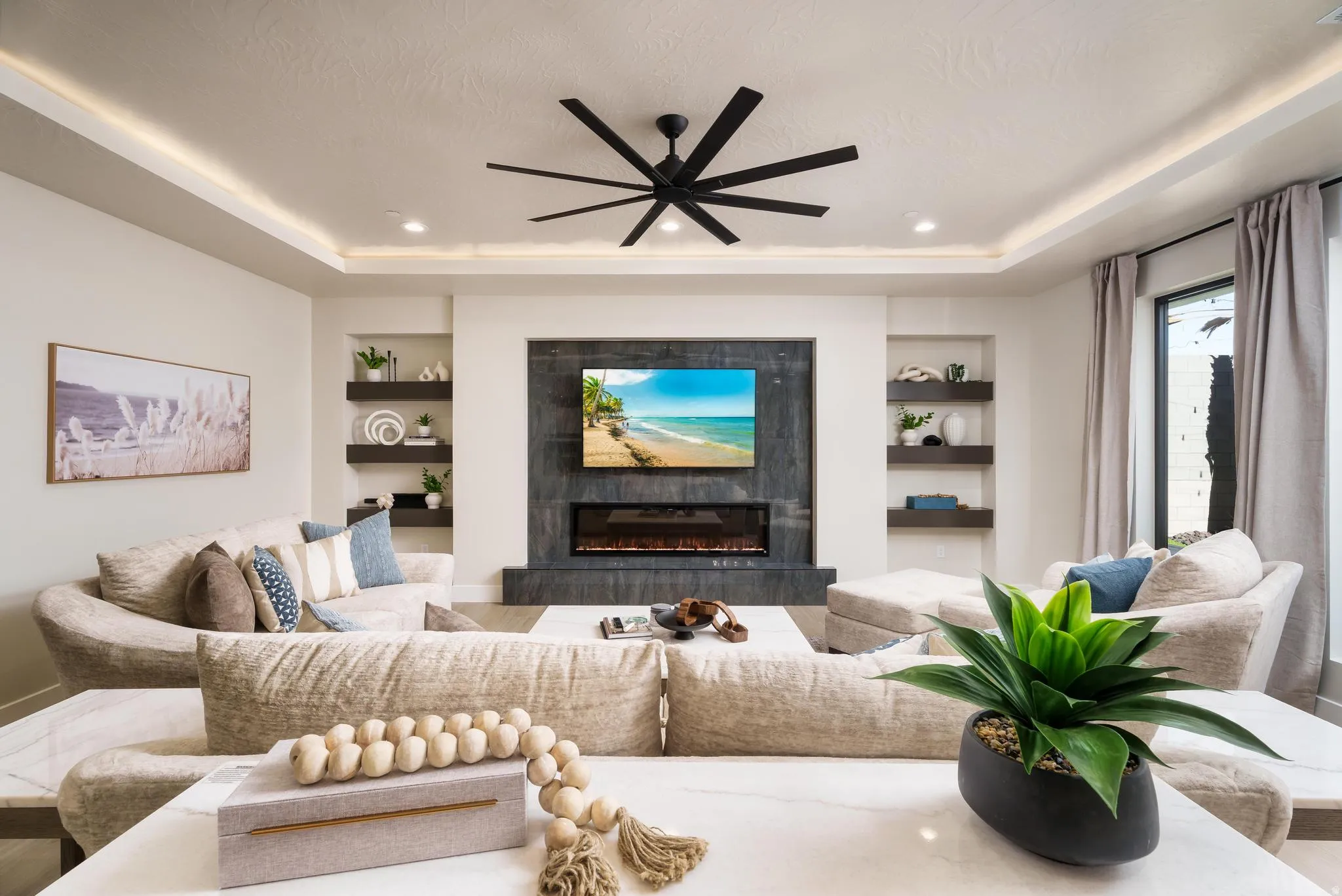 Tray ceiling with recessed lighting and cove lighting, built-in shelving, large window with drapes, wall-mounted television, and an electric fireplace with a dark-toned surround