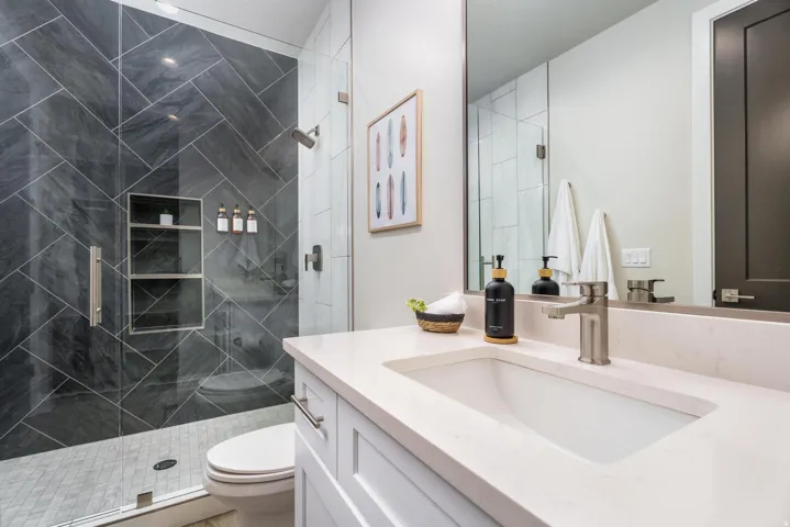 Glass-enclosed shower with dark gray diagonal tile and built-in shelving, white integrated sink, light-colored countertop, and brushed nickel fixtures