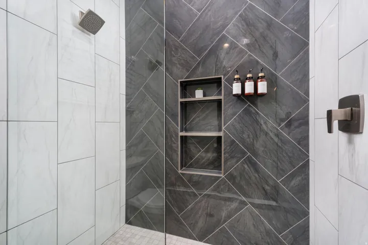 Spa-inspired shower enclosure featuring large-format charcoal tile with diagonal layout, frameless glass partition, dual built-in niches with shelving, and contemporary wall-mounted fixtures