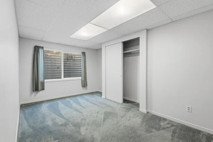 Wall-to-wall carpeting, window with privacy blinds, drop ceiling with integrated lighting, and a closet with sliding doors