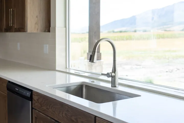 Stainless steel sink with a gooseneck faucet, light-toned countertop, wood-finish cabinetry, and a subway tile backsplash