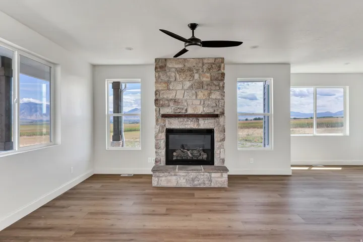 Stone-clad fireplace with a wood mantel, black ceiling fan, wood-finish flooring, and multiple windows