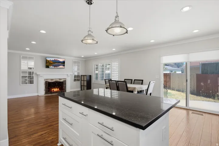 White kitchen island with dark countertop, stainless steel pendant lighting, recessed ceiling lights, wood-finish flooring, and a fireplace with a white mantel