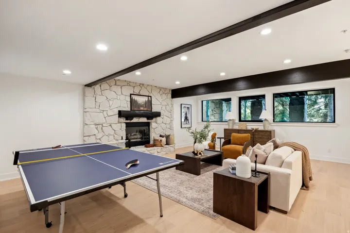 Recessed lighting illuminating a stone fireplace with a dark mantel, wood-finish flooring, and multiple dark wood ceiling beams