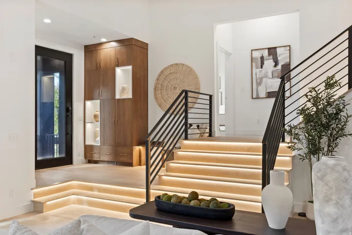 Illuminated staircase with modern horizontal railings, integrated wood-finish cabinetry with display niches, and a full-light entry door