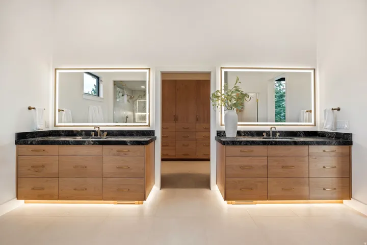 Dual wood-finish vanities with dark countertops, integrated sinks, backlit mirrors, and recessed floor lighting