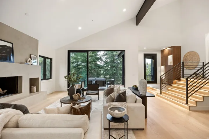 Vaulted ceiling with exposed beam, expansive sliding glass doors, contemporary fireplace with integrated shelving, light wood-finish flooring, and a modern staircase with metal railings