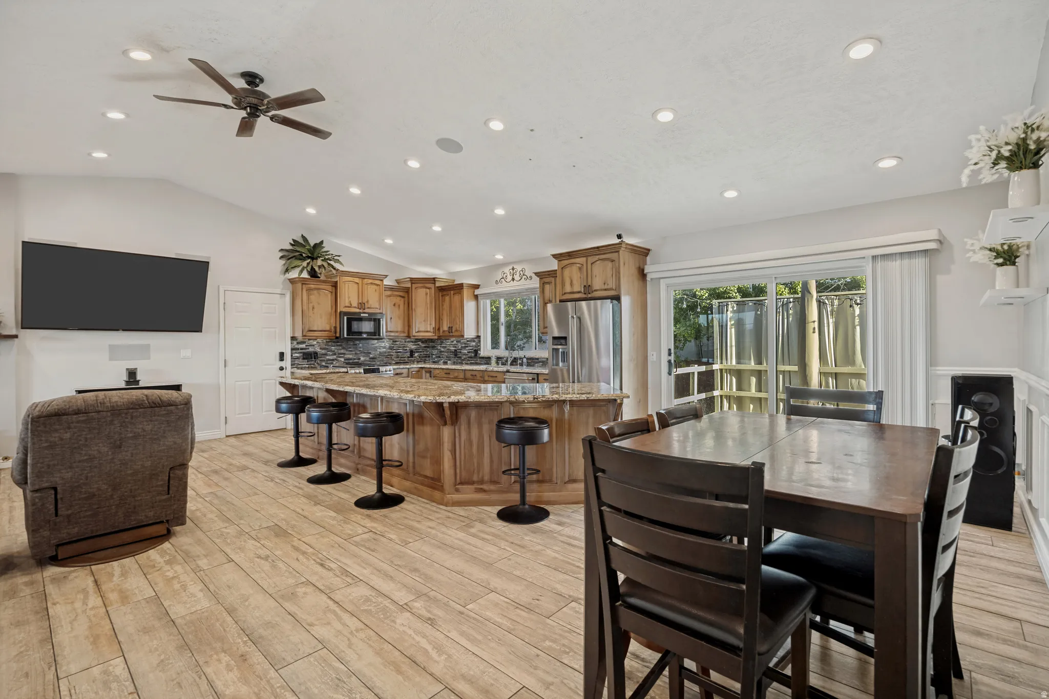 Vaulted ceiling with recessed lighting and a ceiling fan, light wood-finish flooring, wood cabinetry, stone countertops, and sliding glass doors