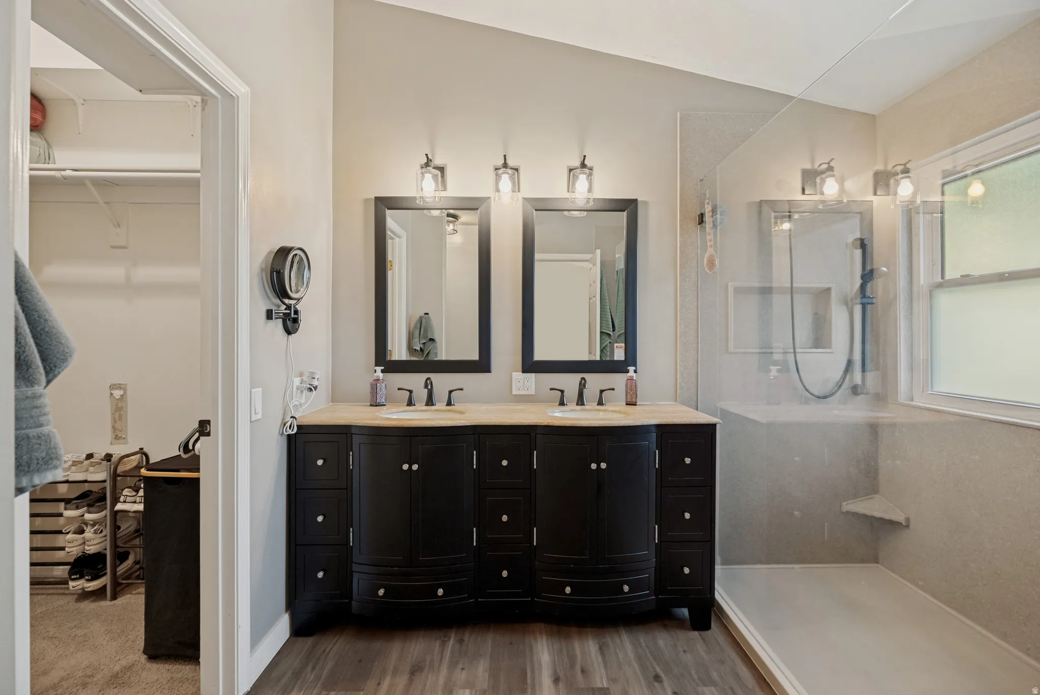 Dual vanity with undermount sinks, wood-finish flooring, glass-enclosed shower, built-in shower niche, and a window for natural light