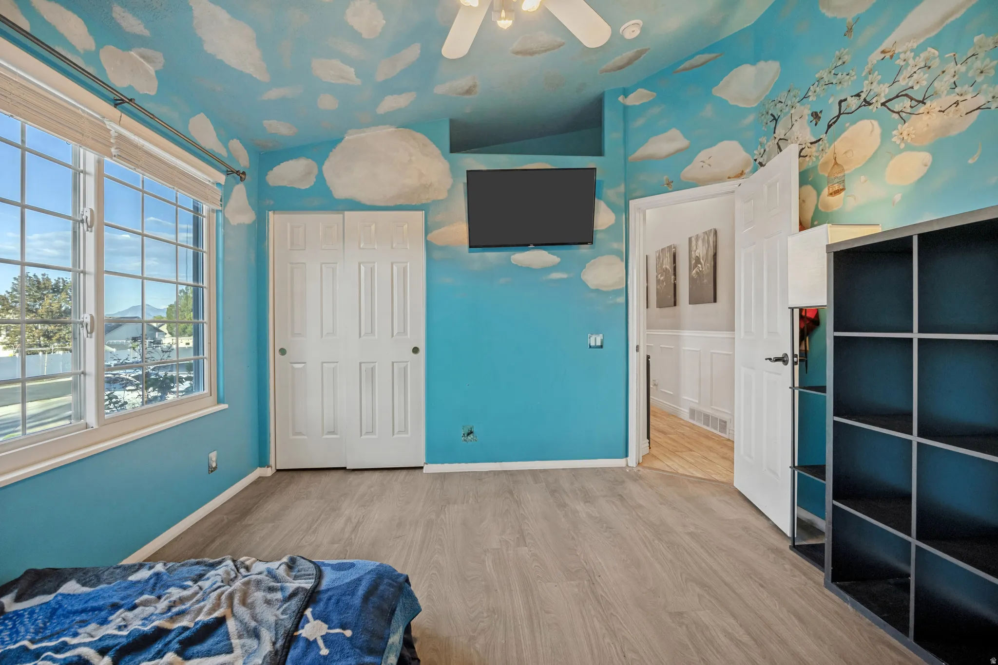 Window with muntins, wood-finish flooring, white bi-fold closet doors, ceiling fan, and painted wall mural