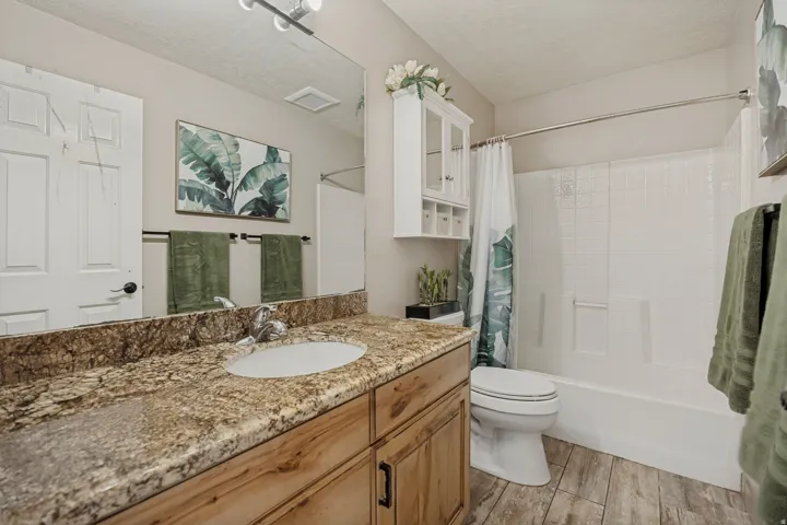 Granite-style countertop with an undermount sink, wood-finish vanity, wood-finish flooring, and a white tub with white tile-style surround