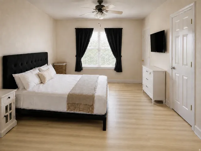 Light-toned wood-finish flooring, ceiling fan with light kit, white six-panel door, window with blinds, and neutral wall tones