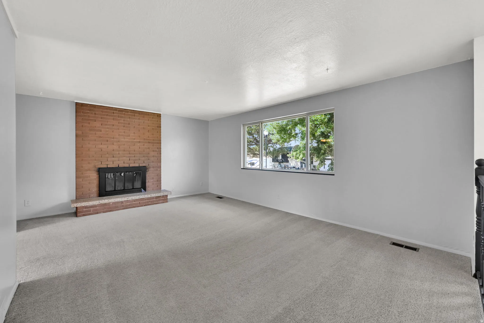 Expansive interior with light gray walls, plush carpeting, a large picture window, and a brick-faced fireplace with a stone hearth