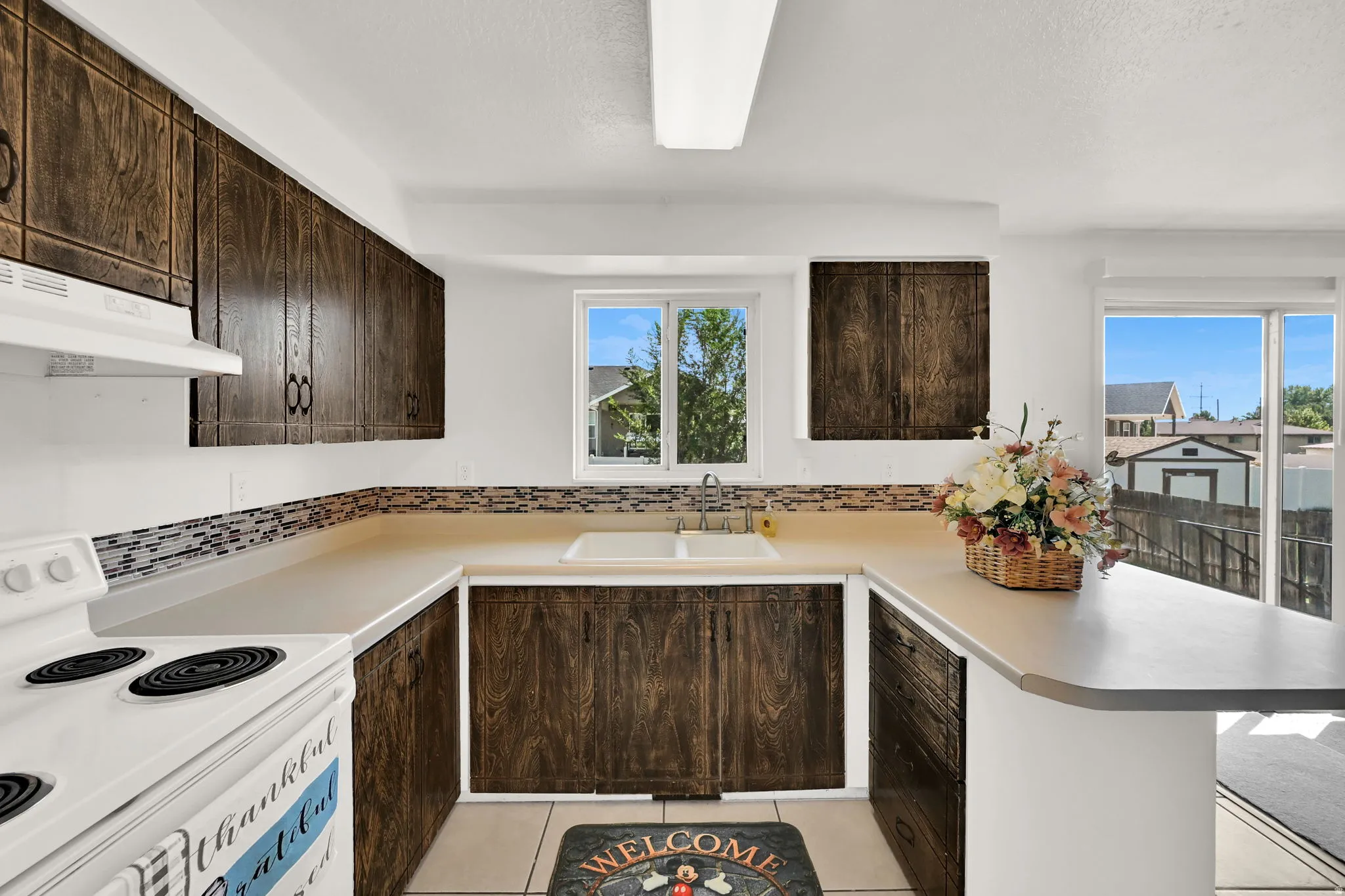 U-shaped kitchen with dark wood-finish cabinetry, a dual-basin sink, mosaic tile backsplash, and light countertops
