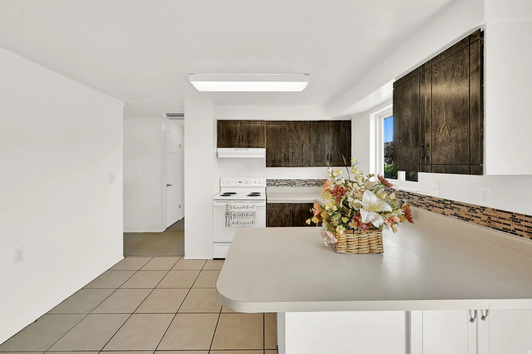 White appliances, dark wood-finish cabinetry, tile flooring, mosaic tile backsplash, and light-colored countertops