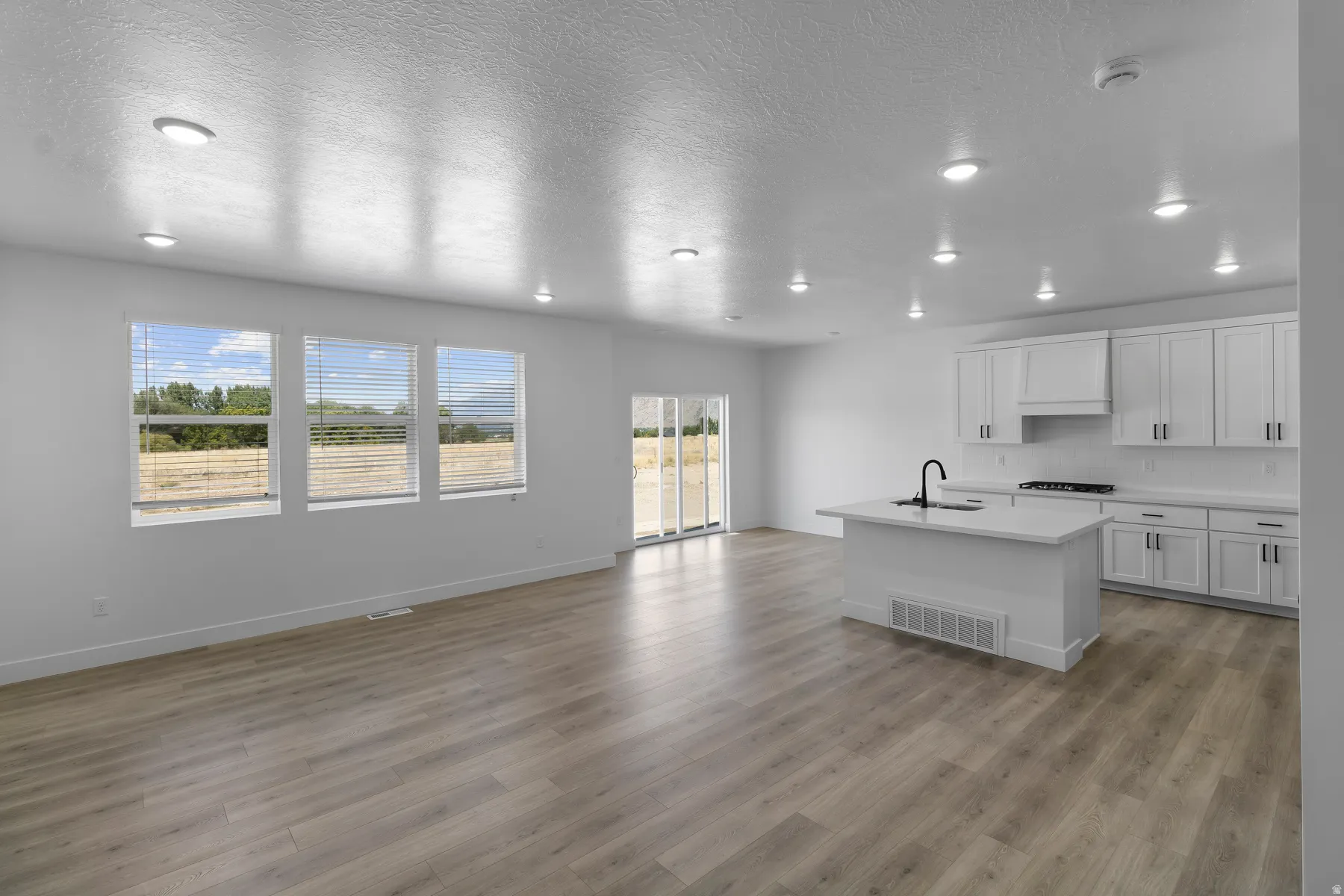 Open-concept living area featuring wood-finish flooring, recessed lighting, and a kitchen with white cabinetry, a center island with a sink, and a sliding glass door