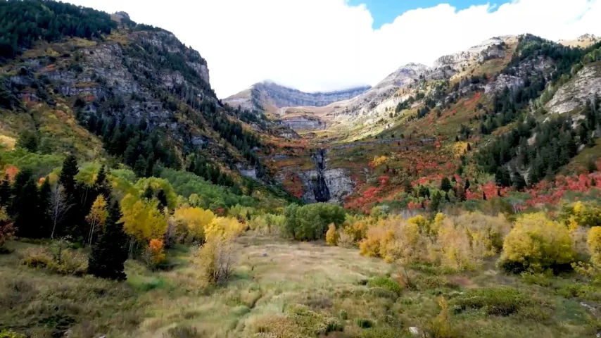 Natural mountain landscape featuring a waterfall, diverse tree foliage, and an expansive grassy terrain