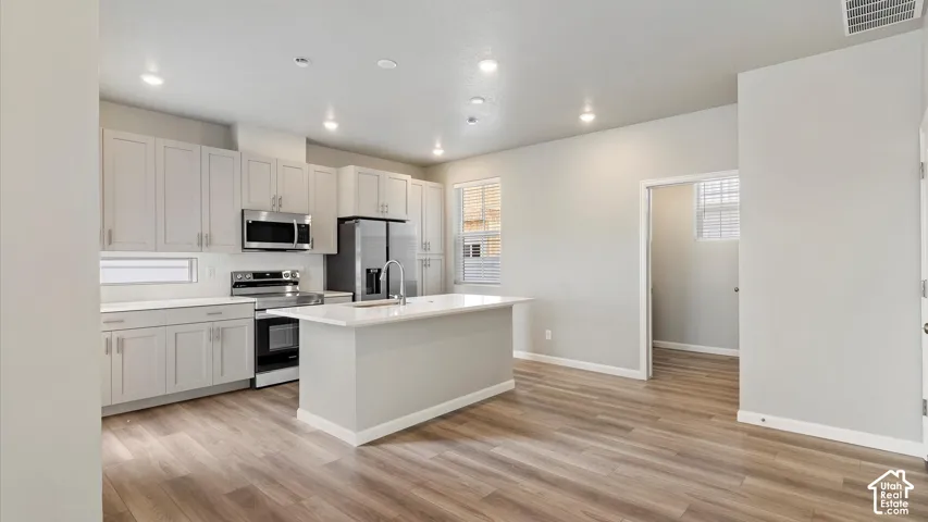 Open-concept kitchen featuring light-toned cabinetry, stainless steel appliances, a center island with sink, and wood-finish flooring