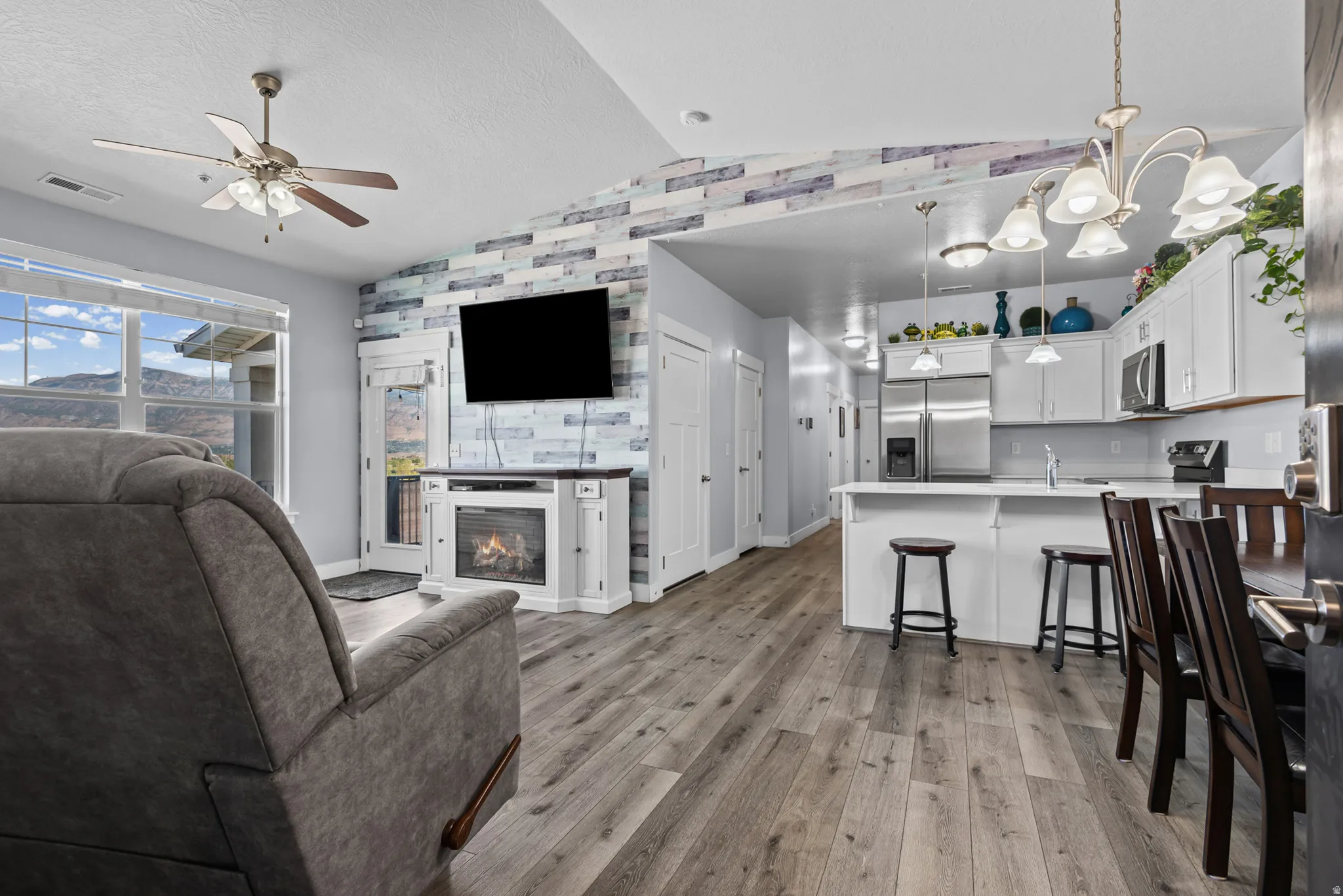 Vaulted ceiling with a ceiling fan, fireplace with a decorative mantel, wood-finish flooring, white kitchen cabinetry, and stainless steel appliances