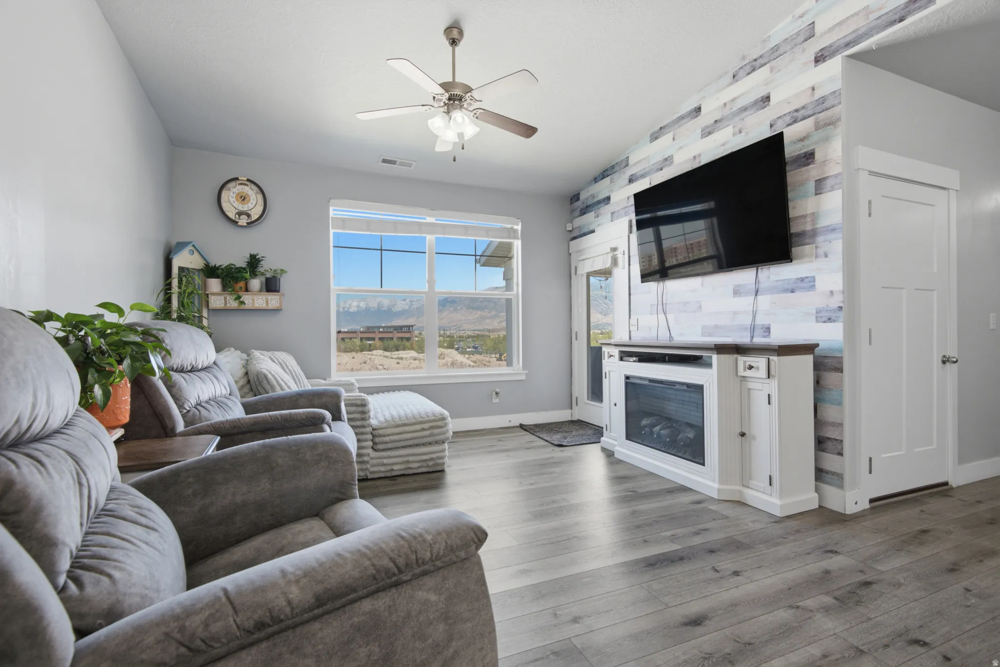 Vaulted ceiling with a ceiling fan, large picture window, wood-finish flooring, accent wall with horizontal paneling, and an electric fireplace
