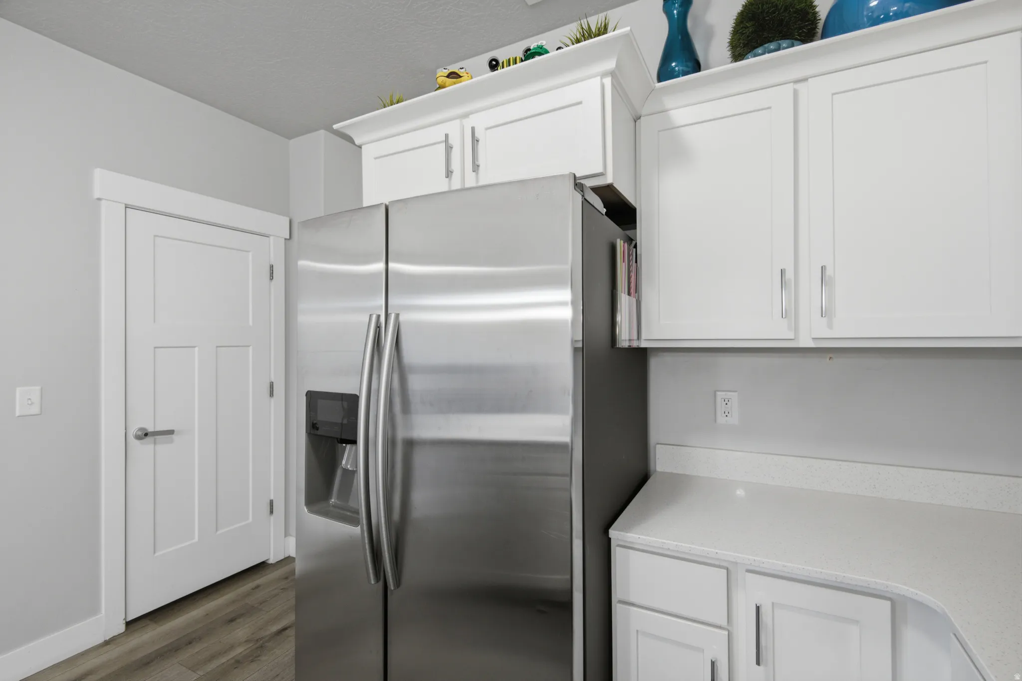 Stainless steel refrigerator with a built-in dispenser, white shaker-style cabinetry, light-toned countertops, and wood-finish flooring