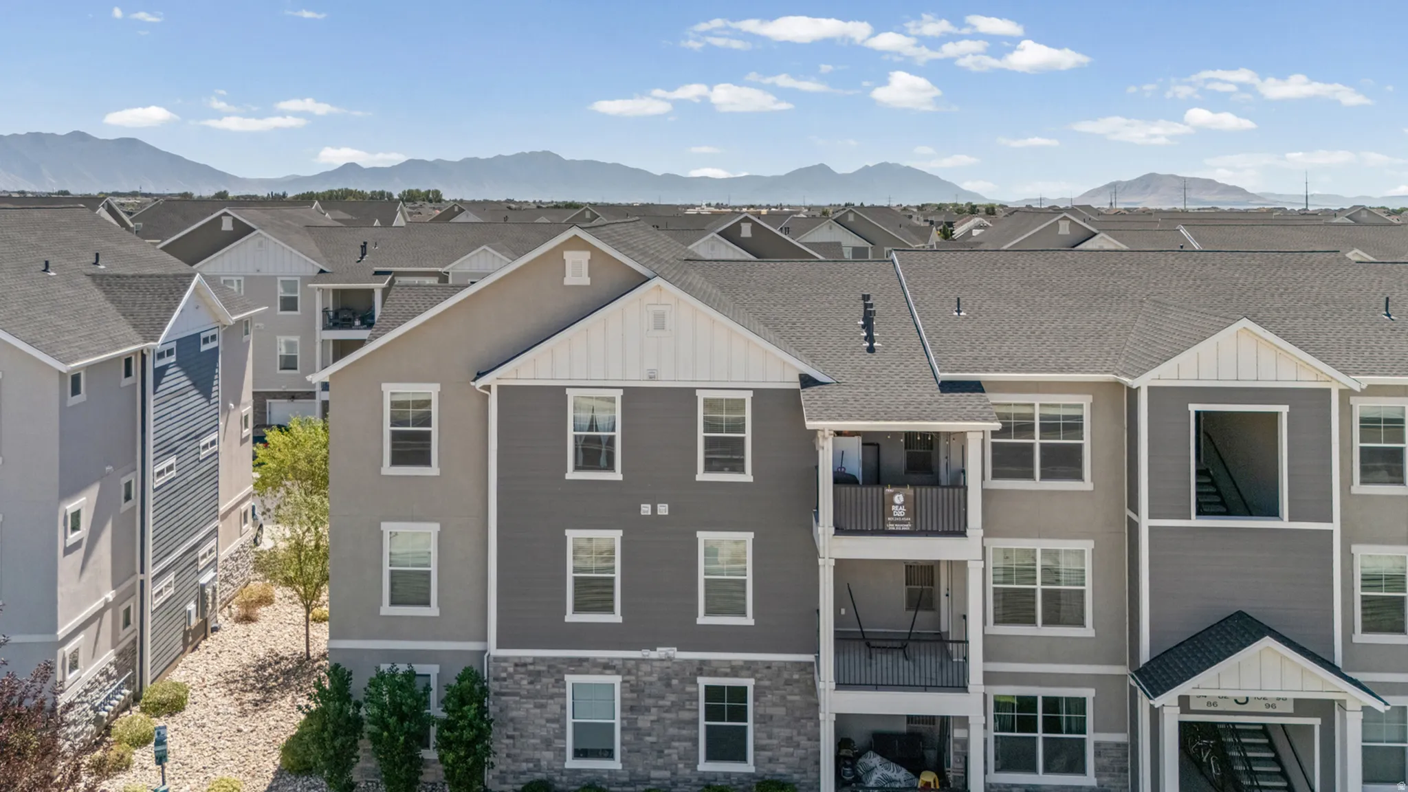 Multi-story residential building featuring balconies with metal railings, a stone facade on the lower level, and mountain scenery in the background