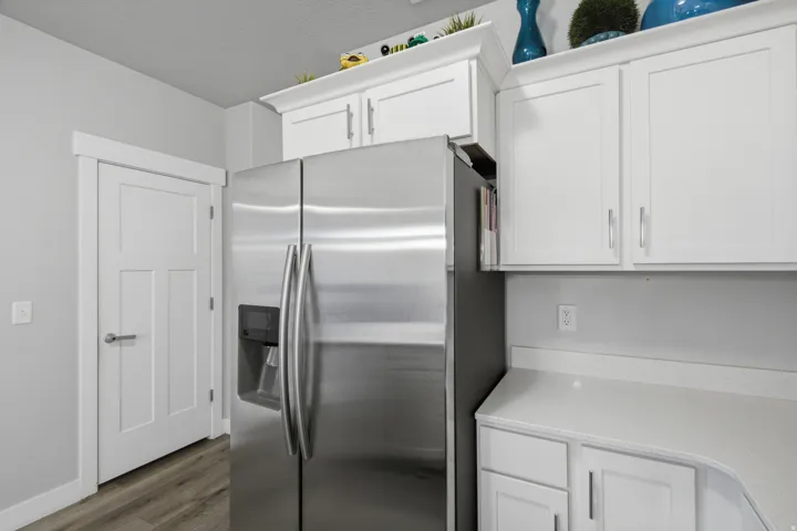 Stainless steel refrigerator with a built-in dispenser, white shaker-style cabinetry, light-toned countertops, and wood-finish flooring