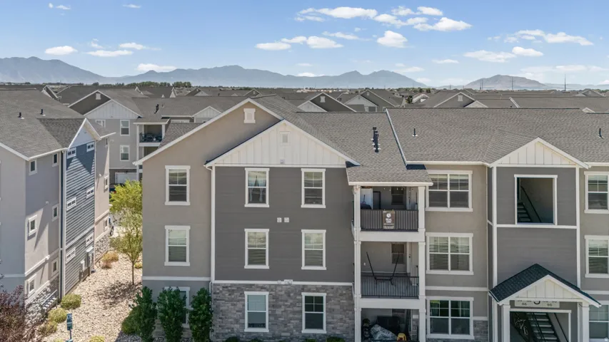 Multi-story residential building featuring balconies with metal railings, a stone facade on the lower level, and mountain scenery in the background