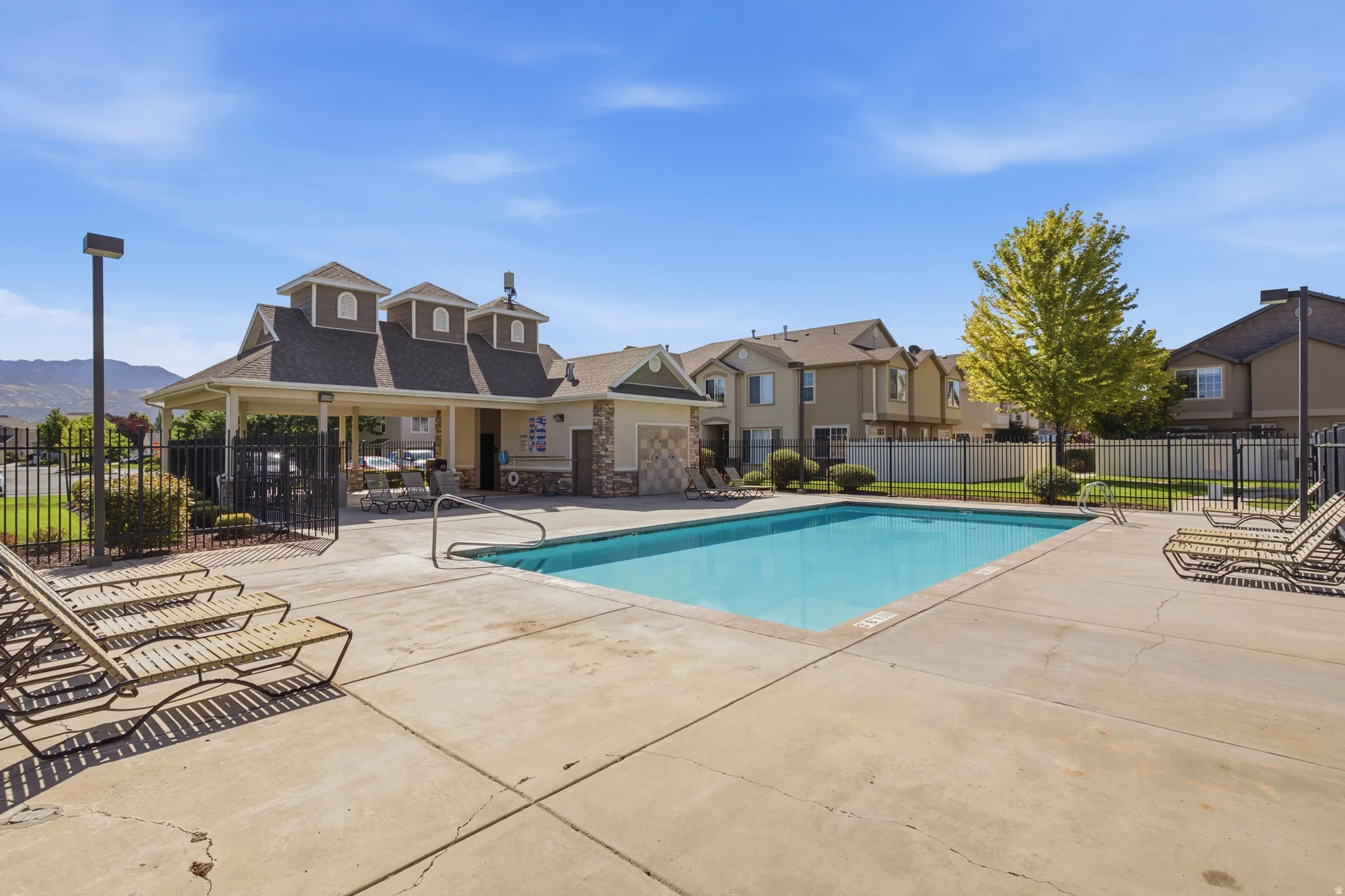 Outdoor community swimming pool with concrete decking, surrounding black metal fencing, and an adjacent covered structure with stone accents