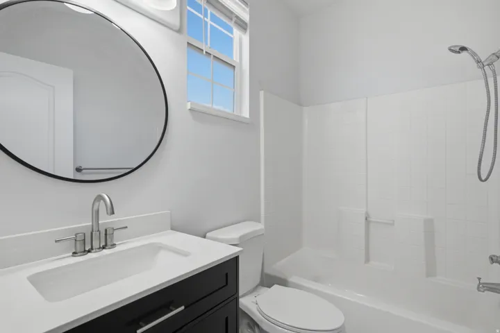 Bathroom featuring a vanity with an integrated sink, a large round mirror, a window with blinds, and a shower/tub combination with a tiled surround