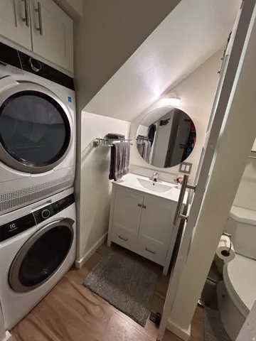 Integrated laundry appliances with upper cabinetry, wood-finish flooring, white vanity with basin sink, circular wall mirror, and modern wall sconce