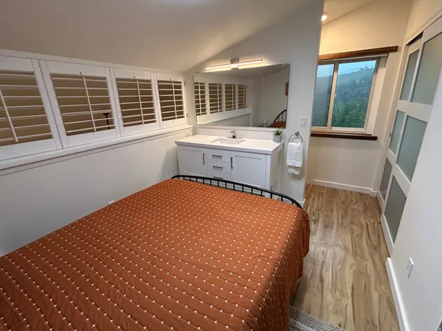 Angled ceiling with track lighting, white vanity with integrated sink, large wall-mounted mirror, white plantation shutters, and wood-finish flooring