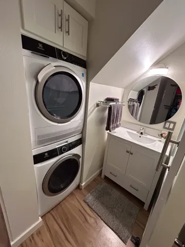 Stacked washer and dryer, wood-finish flooring, white vanity with integrated sink, wall-mounted oval mirror, and built-in upper cabinetry