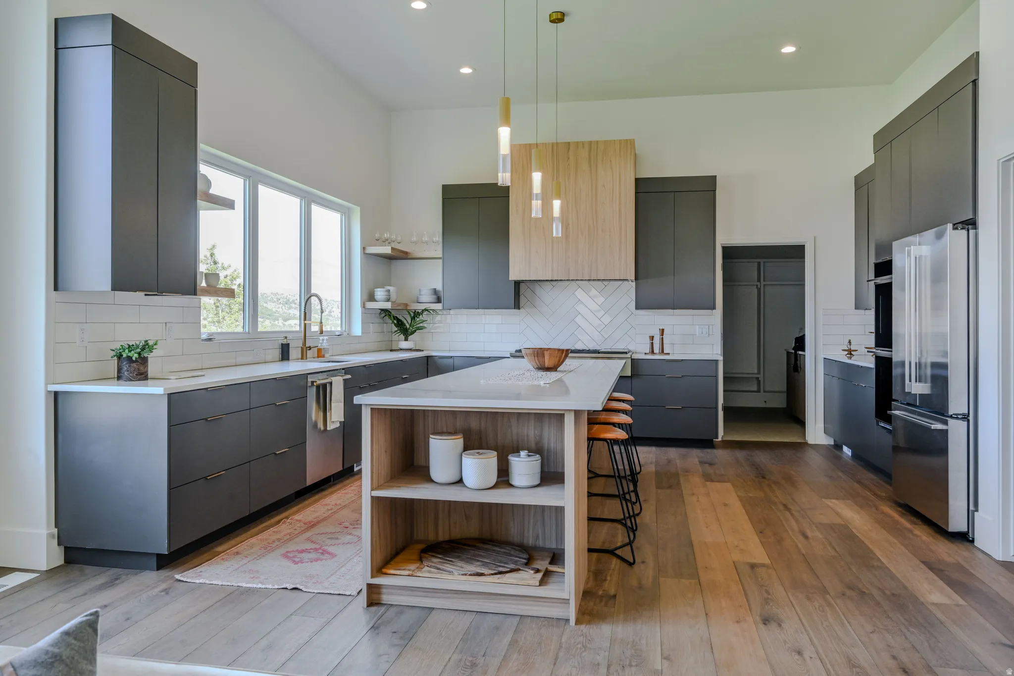 Modern kitchen with wood-finish flooring, dark cabinetry, a central island with integrated shelving, stainless steel appliances, and a white subway tile backsplash
