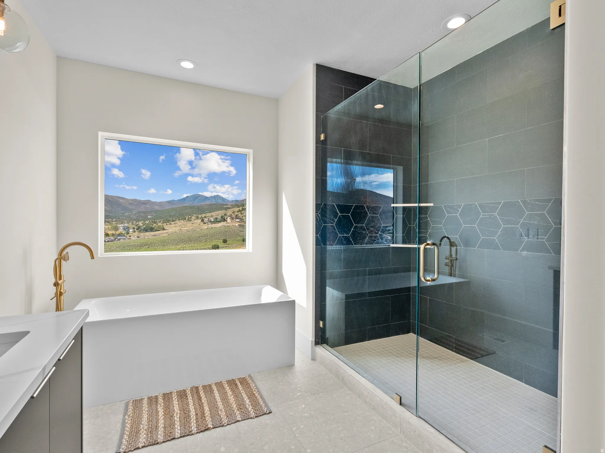 Freestanding soaking tub with gold-finish fixture, expansive window with mountain scenery, glass-enclosed shower with dark tile surround, integrated shower bench, and hexagonal accent tiling