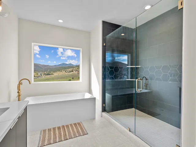 Freestanding soaking tub with gold-finish fixture, expansive window with mountain scenery, glass-enclosed shower with dark tile surround, integrated shower bench, and hexagonal accent tiling